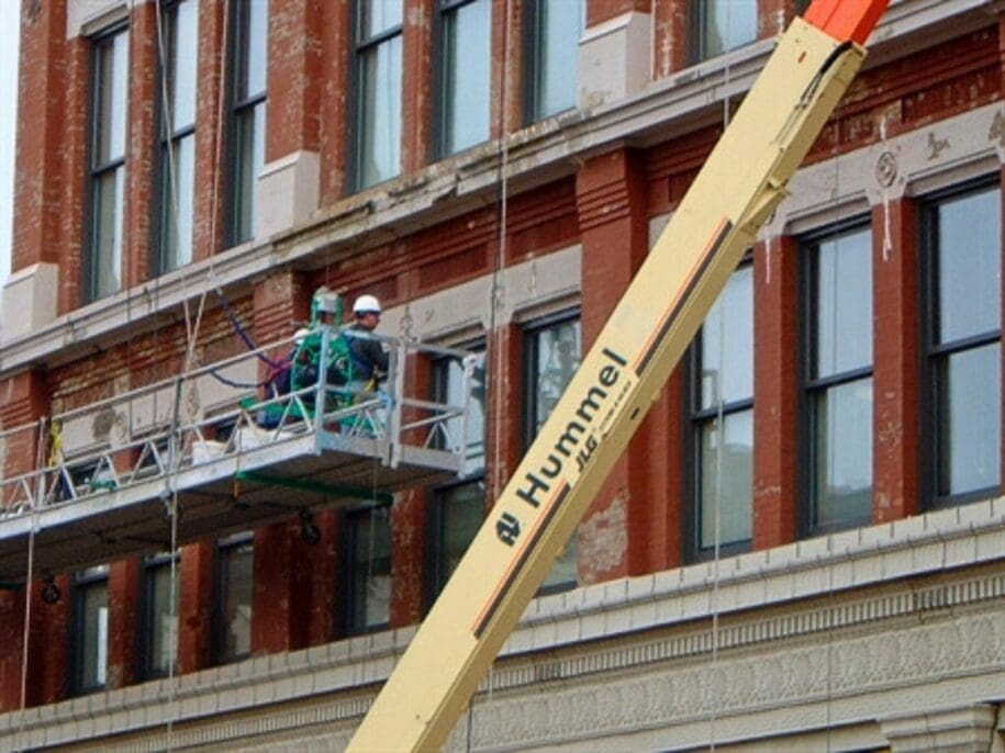 Two workers on suspended platform performing masonry facade repair on brick building in Cincinnati, Ohio, with Hummel crane visible