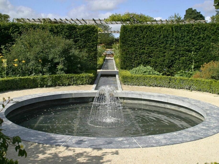 Circular stone fountain with central water jet in formal garden surrounded by manicured hedges and pergola