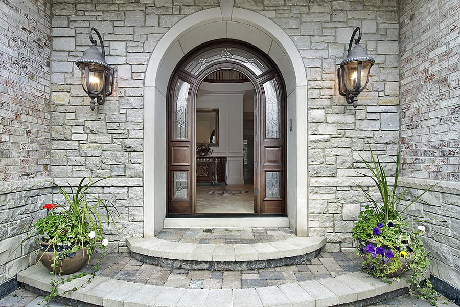 Elegant arched entryway with light stone facade, dark wooden double doors with glass panels, flanked by wall sconces and flowering planters on a curved stone landing