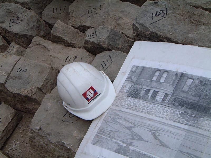 White hard hat with red logo resting on stone blocks marked with numbers, blueprint of brick building facade visible beside rocks on ground